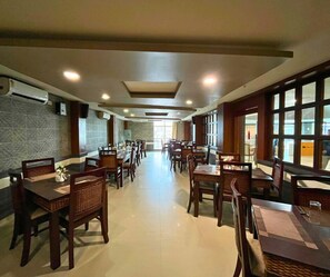 A spacious dining area with multiple tables and chairs, a stone wall on one side, and large windows on the other. - Hotel Pybss (Papumpare)