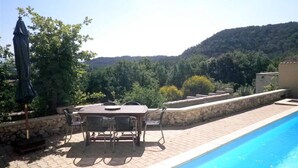 An outdoor pool area with a table and chairs, surrounded by a stone wall and a view of a forested hillside. - Villa with private pool, views of the Ventoux (Saint Paul trois Chateaux)