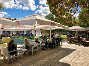 An outdoor seating area with tables and chairs under white umbrellas, a pool, and people dining and socializing. - Nomado Hostel (Varna)