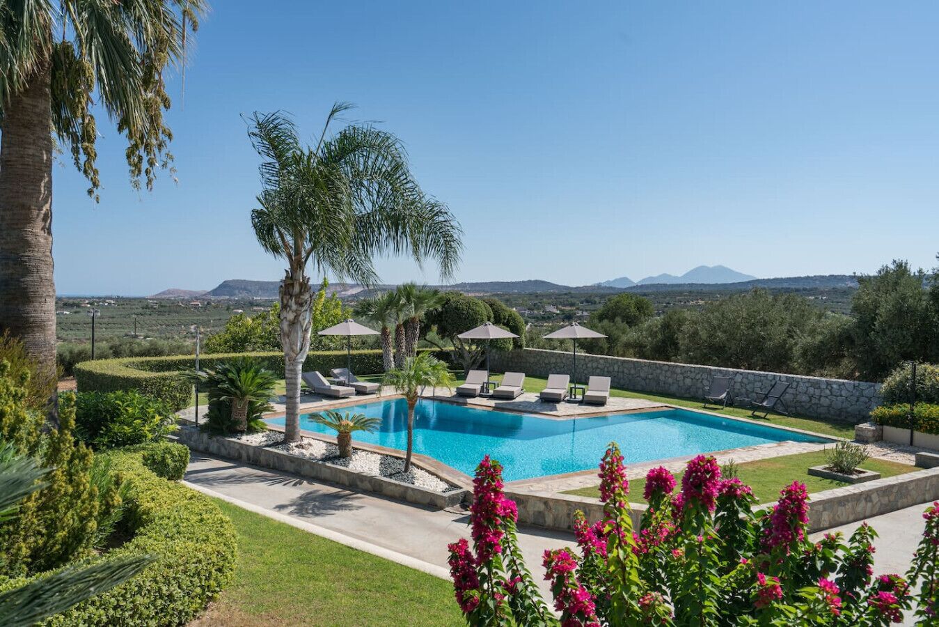 A swimming pool with lounge chairs and umbrellas, surrounded by greenery and a stone wall.