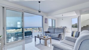 Ampla sala de estar com uma grande porta de vidro deslizante que dá para uma varanda com vista para o mar. Há um sofá cinza, uma mesa de centro e uma escadaria ao fundo.