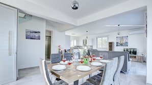 Uma área de refeições moderna, com uma mesa de madeira posta para quatro pessoas, cadeiras estofadas em cinza e uma cozinha ao fundo.