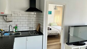 Une cuisine dotée d’une hotte noire, d’un évier avec un robinet et d’un plan de travail sur lequel se trouvent un égouttoir et des produits de nettoyage. Un téléviseur est fixé sur un meuble blanc.