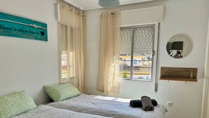 Une chambre avec deux lits, une fenêtre avec des stores, un miroir fixé au mur et une pancarte sur laquelle est écrit : « On est chez soi là où règne l’amour ».