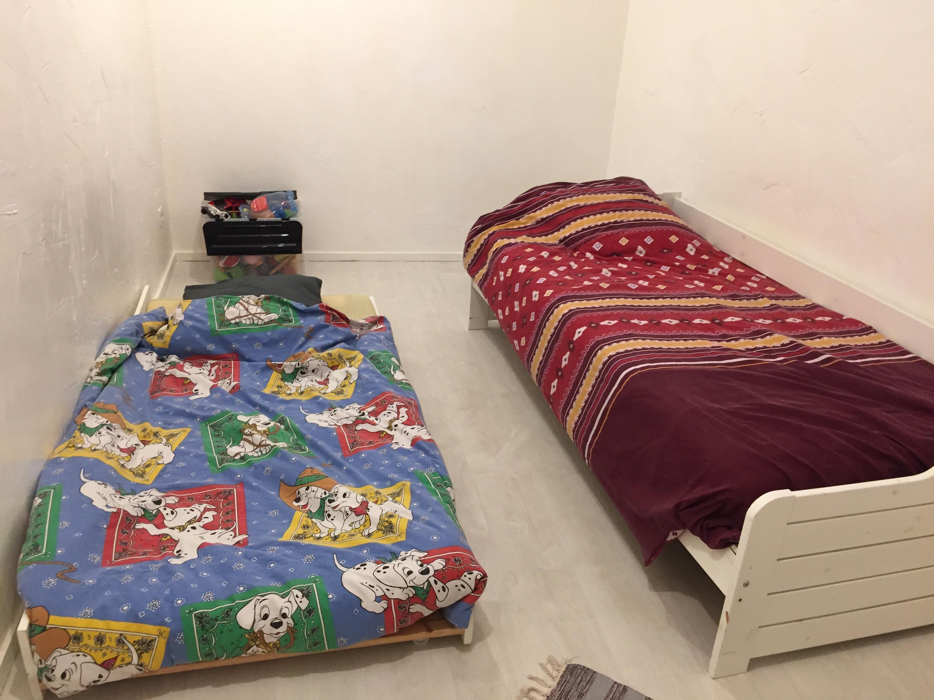 A small, single-bed room with a blue bedspread featuring cartoon characters and a red bedspread with a geometric pattern.