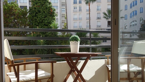 En balkon med træstole og et bord, en plantekasse og udsigt til bygninger og træer.