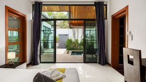 A modern hotel room with a large sliding glass door leading to an outdoor pool area, a bed with neatly arranged pillows, and a small table with a lamp. - Emotion 1 - 3 Bedrooms, Rawai - Phuket (Rawai)