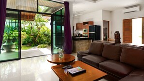 A modern living room with a sofa, a coffee table, and a view of a lush garden through large sliding glass doors. - Emotion 1 - 3 Bedrooms, Rawai - Phuket (Rawai)