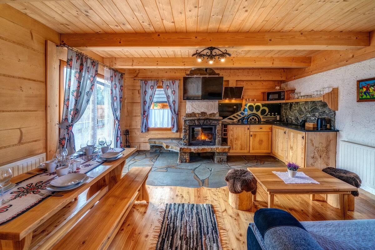 A wooden cabin interior with a stone fireplace, wooden dining table, benches, and a cozy seating area.