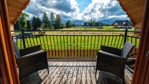 A wooden deck with wicker chairs, a view of a green field, and houses in the background. - Domki Ze Hej z kominkiem lub sauną (Zakopane)