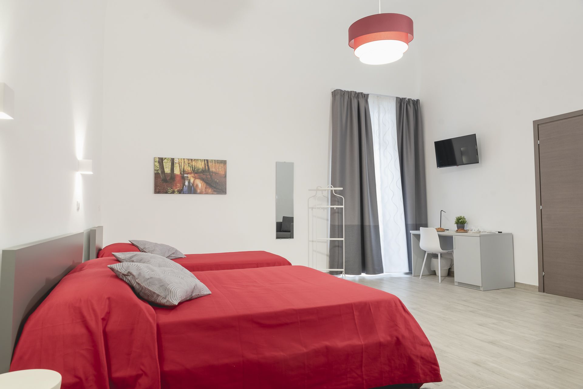 A modern hotel room with two red beds, a desk with a chair, a TV, and a large window with curtains.