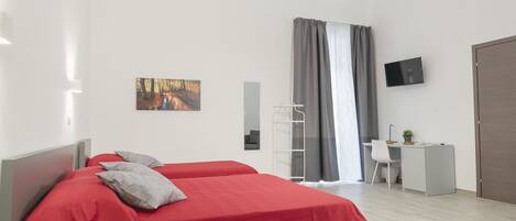 A modern hotel room with two red beds, a desk with a chair, a TV, and a large window with curtains.