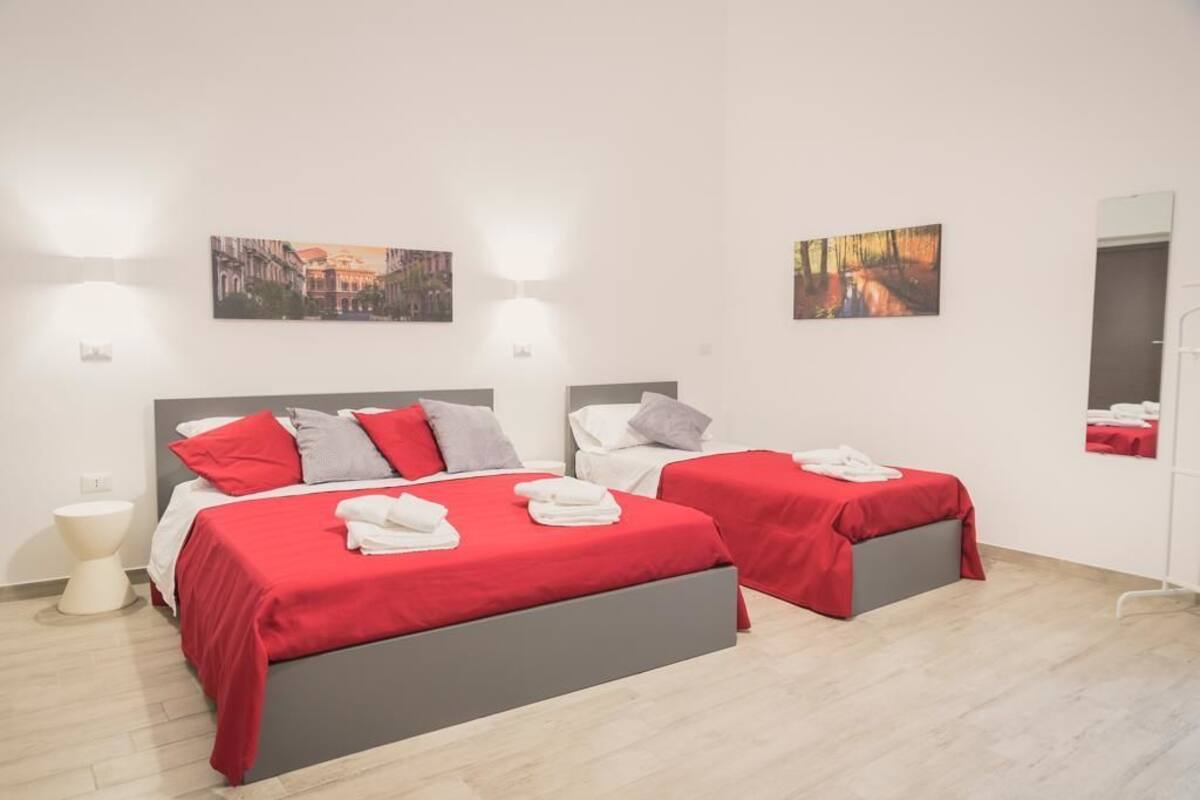 A hotel room with two beds, each with red bedding and white towels. There are two wall-mounted light fixtures and two framed pictures on the walls.