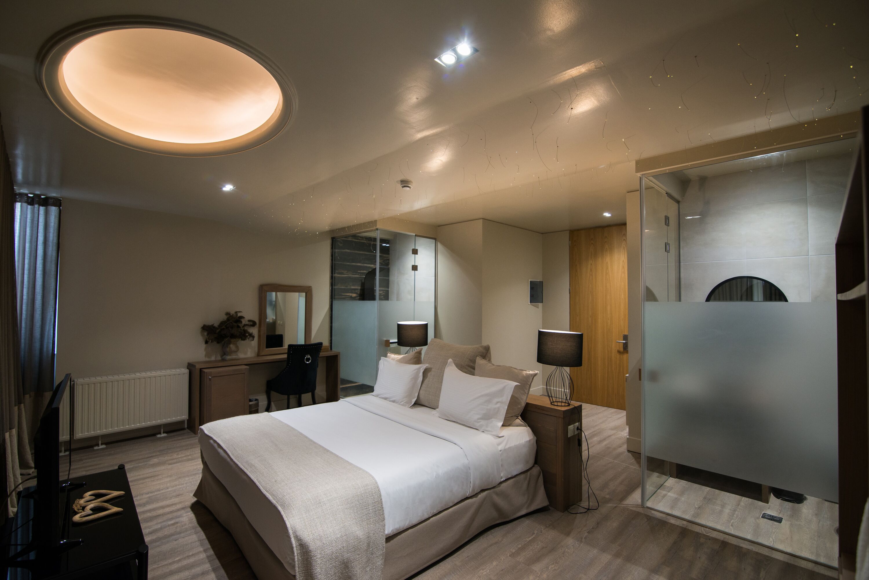 A modern hotel room with a large bed, bedside lamps, a television, and a glass partition wall.