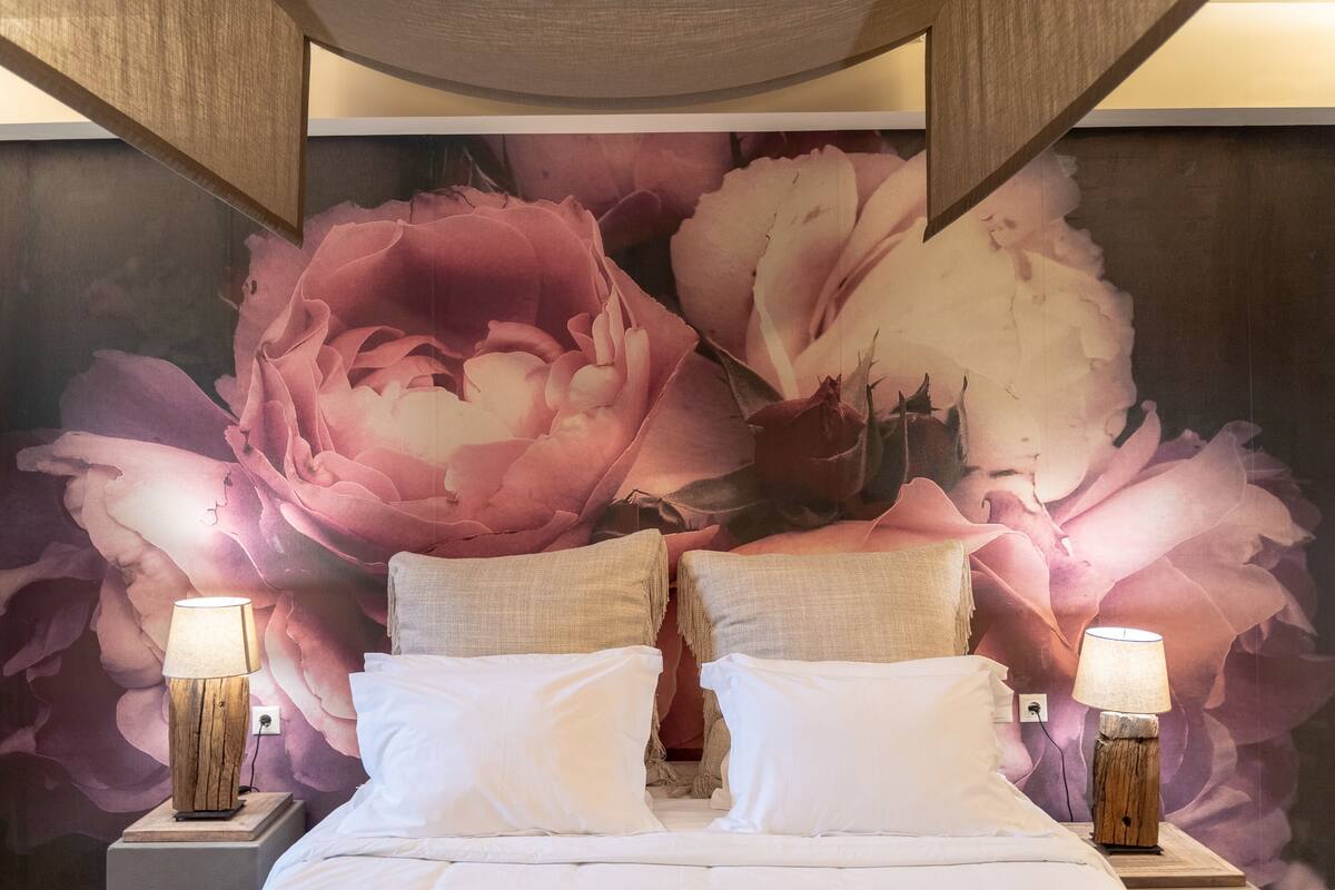 Habitación de hotel con un gran mural floral en la pared, una cama con ropa blanca, dos lámparas de noche y una mesita de noche de madera.
