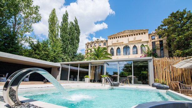 A swimming pool with a modern water feature, surrounded by a wooden deck and a building with arched windows. - Mikelina Boutique Hotel (Pallini)