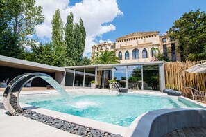 A swimming pool with a modern water feature, surrounded by a wooden deck and a building with arched windows. - Mikelina Boutique Hotel (Pallini)
