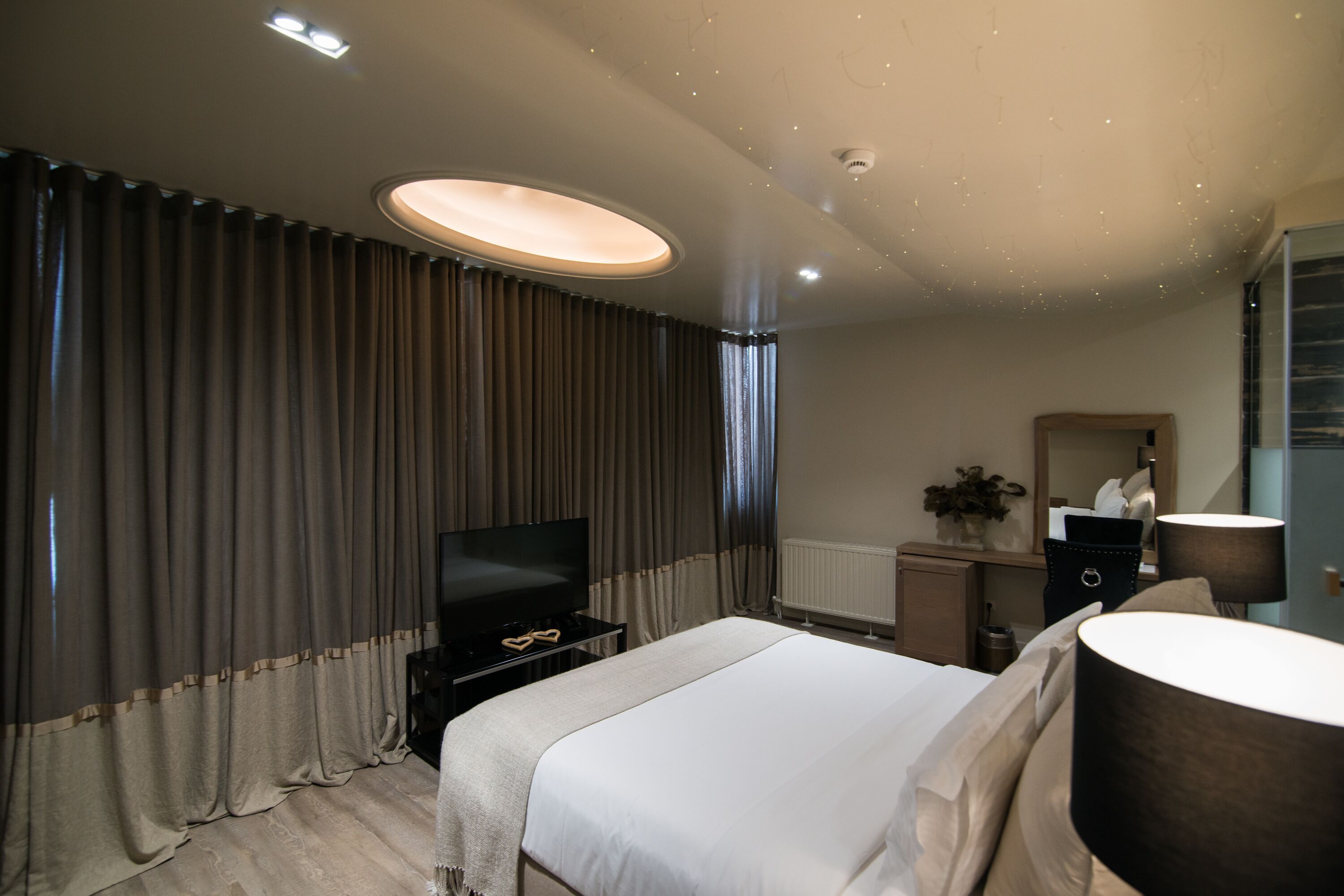 A hotel room with a large bed, a flat-screen TV, a bedside table, a lamp, and a mirror.
