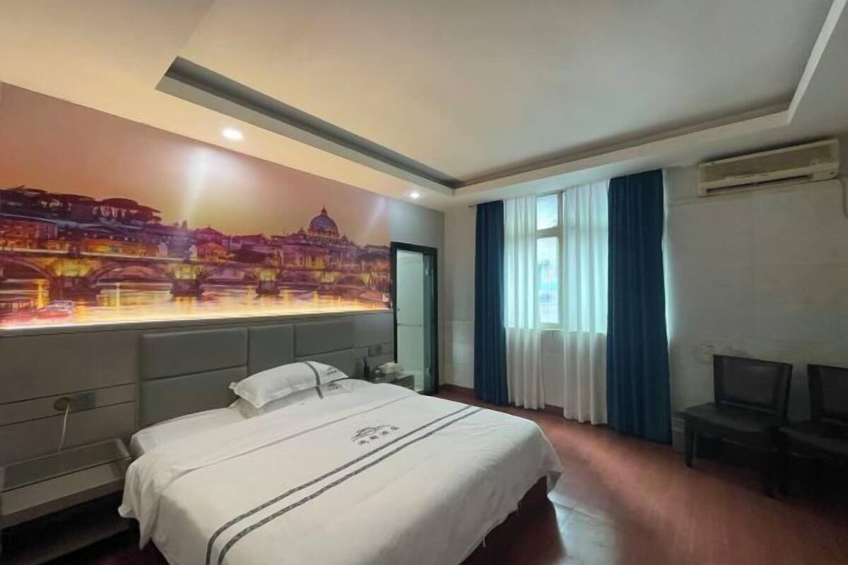 Een hotelkamer met een groot bed, een nachtkastje, een bureau en een stoel. Op de muur is een muurschildering te zien met een stadsbeeld.