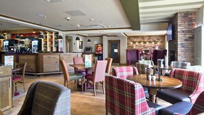 A bar area with a counter, seating with plaid upholstery, and a brick wall. - Premier Inn Cockermouth (Cockermouth)
