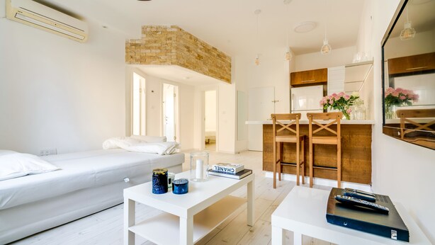 A modern, well-lit room with a white sofa, a small table with books and candles, a kitchen area with wooden chairs, and a brick accent wall. - Double O apartment - Near the bech (Tel Aviv)