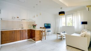 A modern kitchen with wooden cabinets, a white countertop, and a dining area with a white table and chairs. There is a TV mounted on the wall and a window with curtains. - Double O apartment - Near the bech (Tel Aviv)