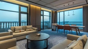 A modern hotel room with a sofa, a round coffee table, a dining area with chairs, and large windows offering a view of the sea. - Weihai Dongshan Hotel (Weihai)