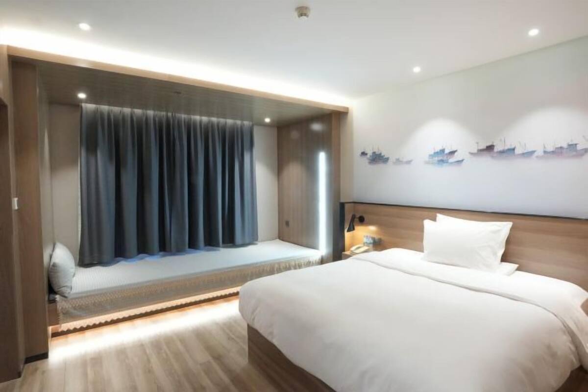 Une chambre d’hôtel comprenant un lit, un coin salon avec des rideaux, une décoration murale représentant un navire, et une table de chevet avec une lampe.