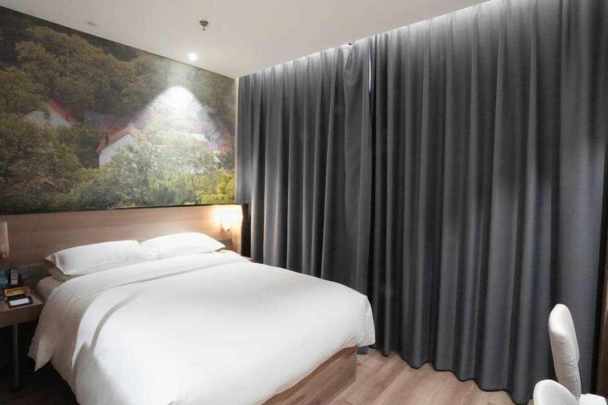Une chambre d’hôtel avec un lit, une table de chevet, une lampe fixée au mur et une grande fenêtre avec des rideaux.