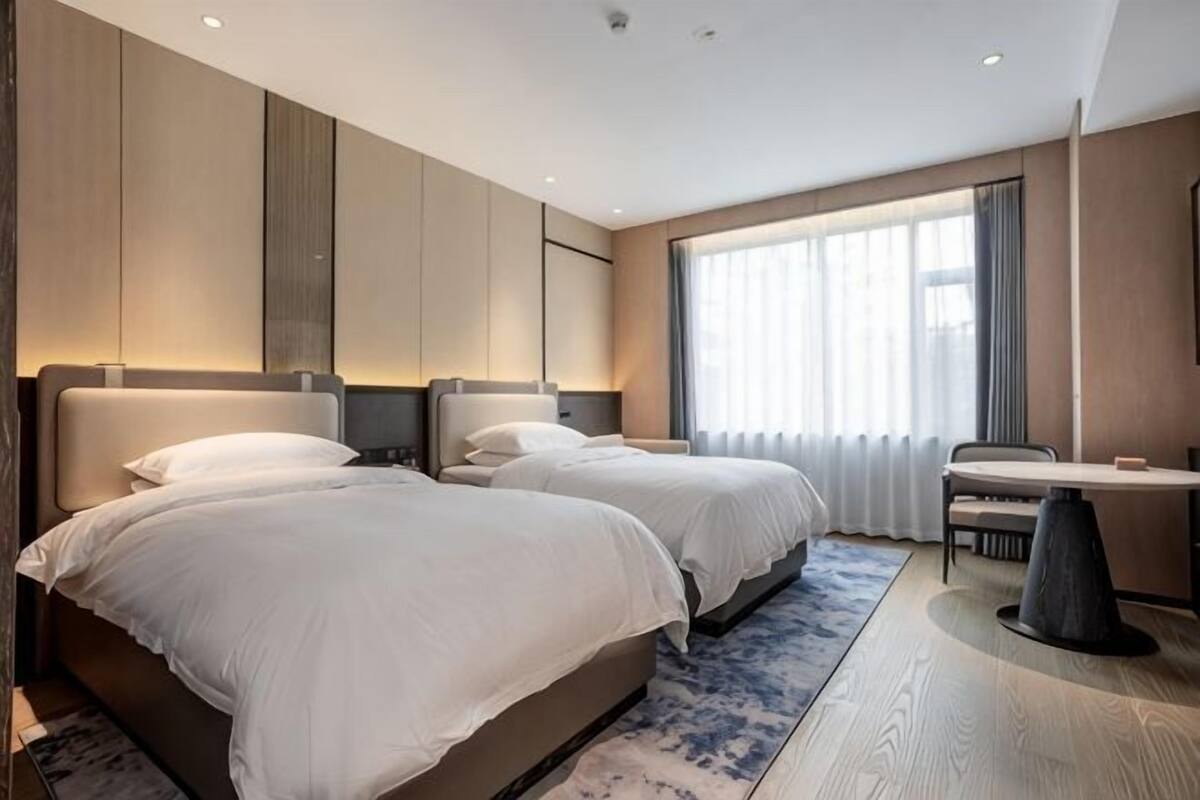 Une chambre d’hôtel avec deux lits, une petite table et une grande fenêtre avec des rideaux.
