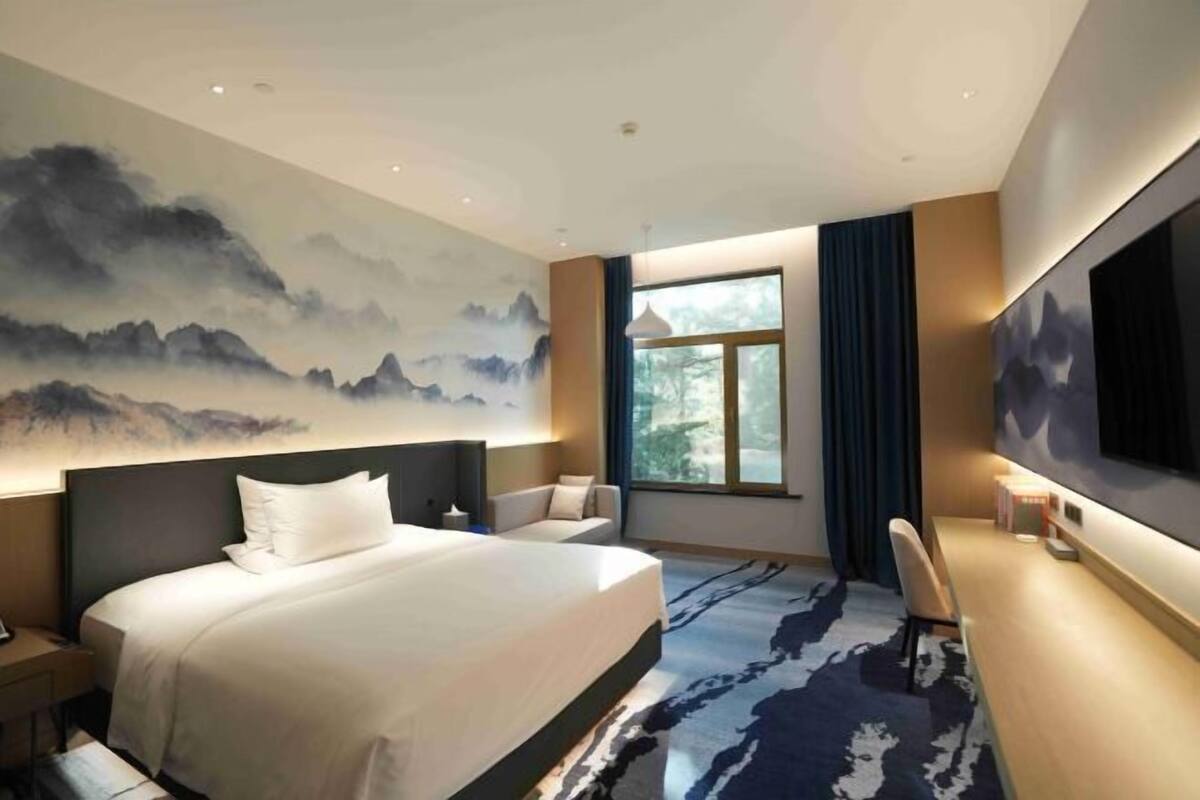 Une chambre d’hôtel avec un lit, un bureau, une chaise, une télévision, une fenêtre et un tableau au mur.