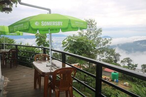 Relax on the deck with a table and two chairs, shaded by a green umbrella, overlooking a lush landscape and distant mountains.