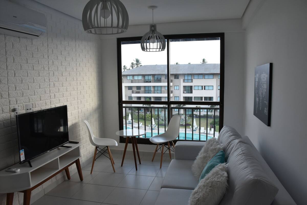 A modern living room with a flat-screen TV, a white sofa, a dining table, and chairs. A large window offers a view of a building and a pool.
