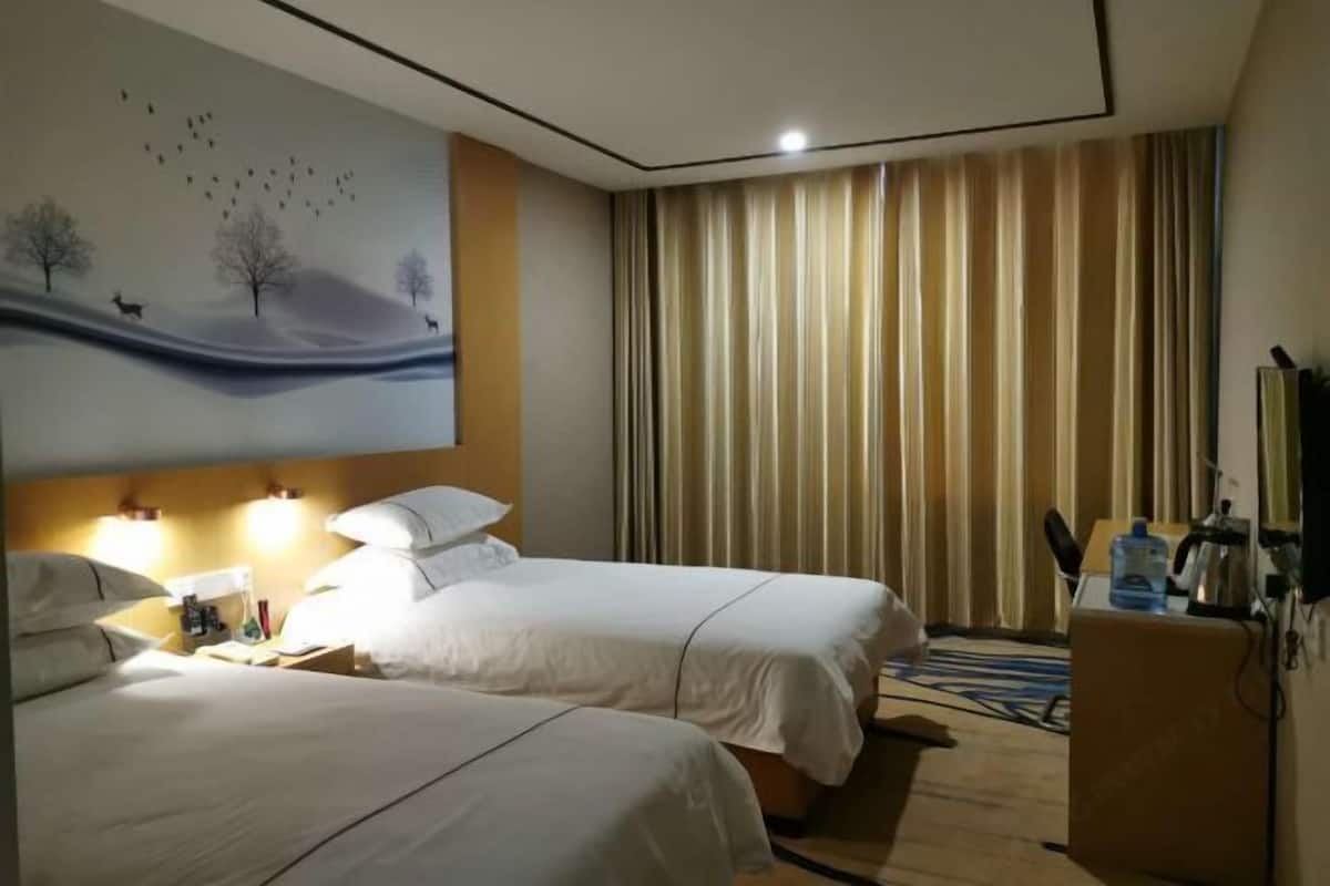 Une chambre d’hôtel avec deux lits, un tableau représentant un paysage, un bureau avec une lampe et une grande fenêtre avec des rideaux.