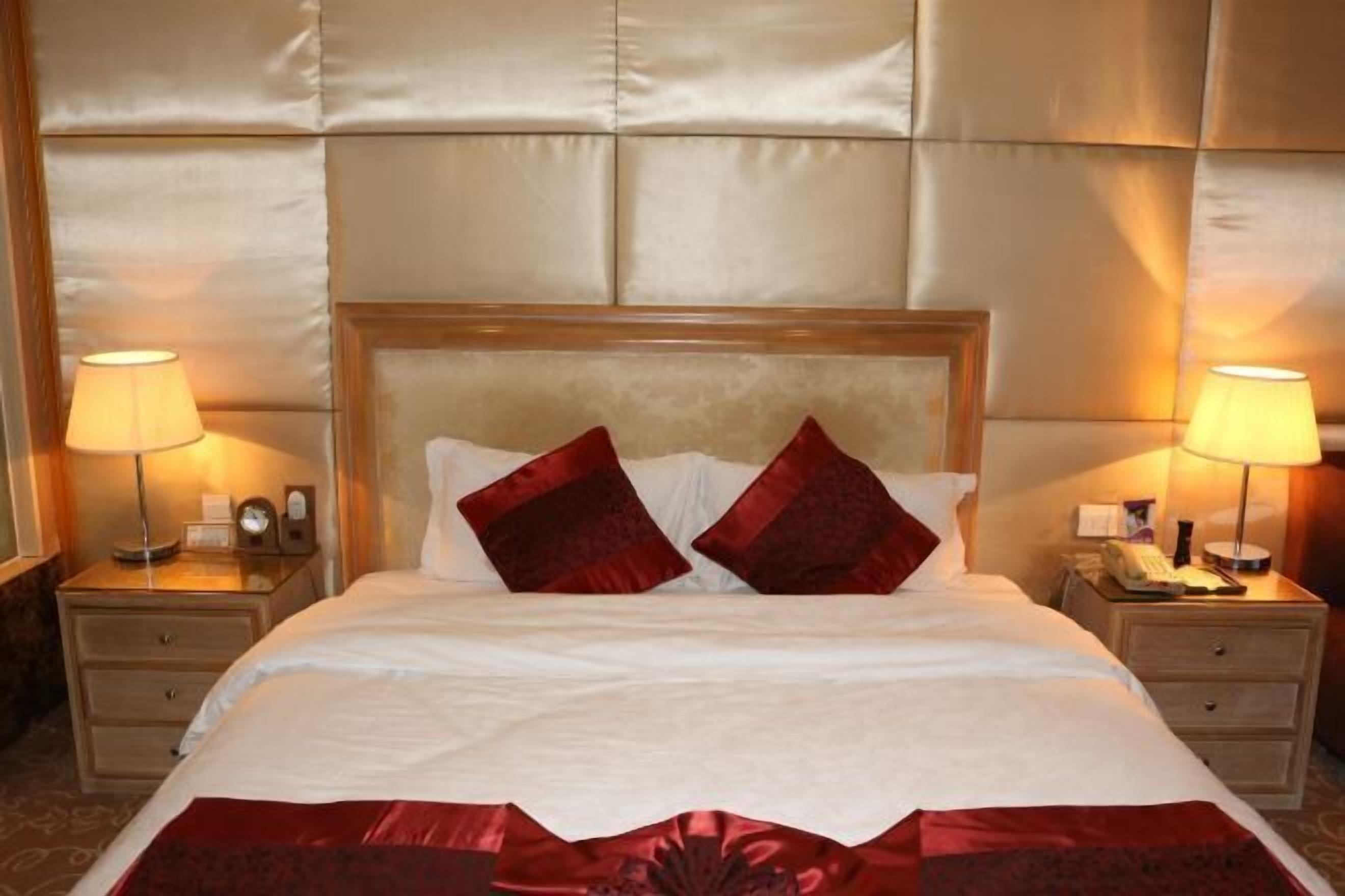 Quarto de hotel com uma cama grande, duas mesas de cabeceira com luminárias, uma cabeceira de madeira e um travesseiro decorativo vermelho.