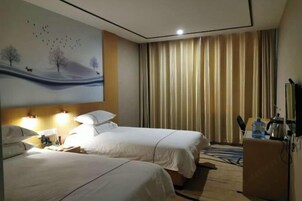 Habitación de hotel con dos camas, un cuadro de un paisaje, un escritorio con una lámpara y vistas al exterior a través de cortinas.