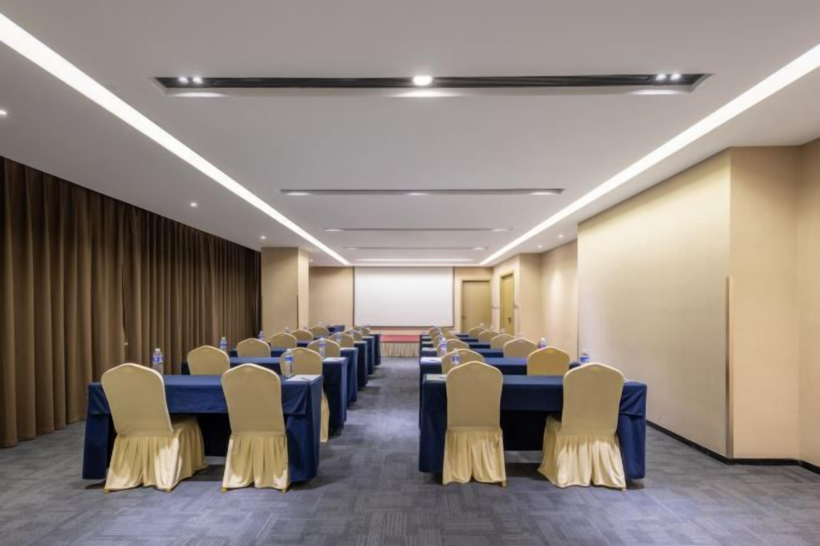 A conference room with rows of tables and chairs, a projector screen, and a carpeted floor.