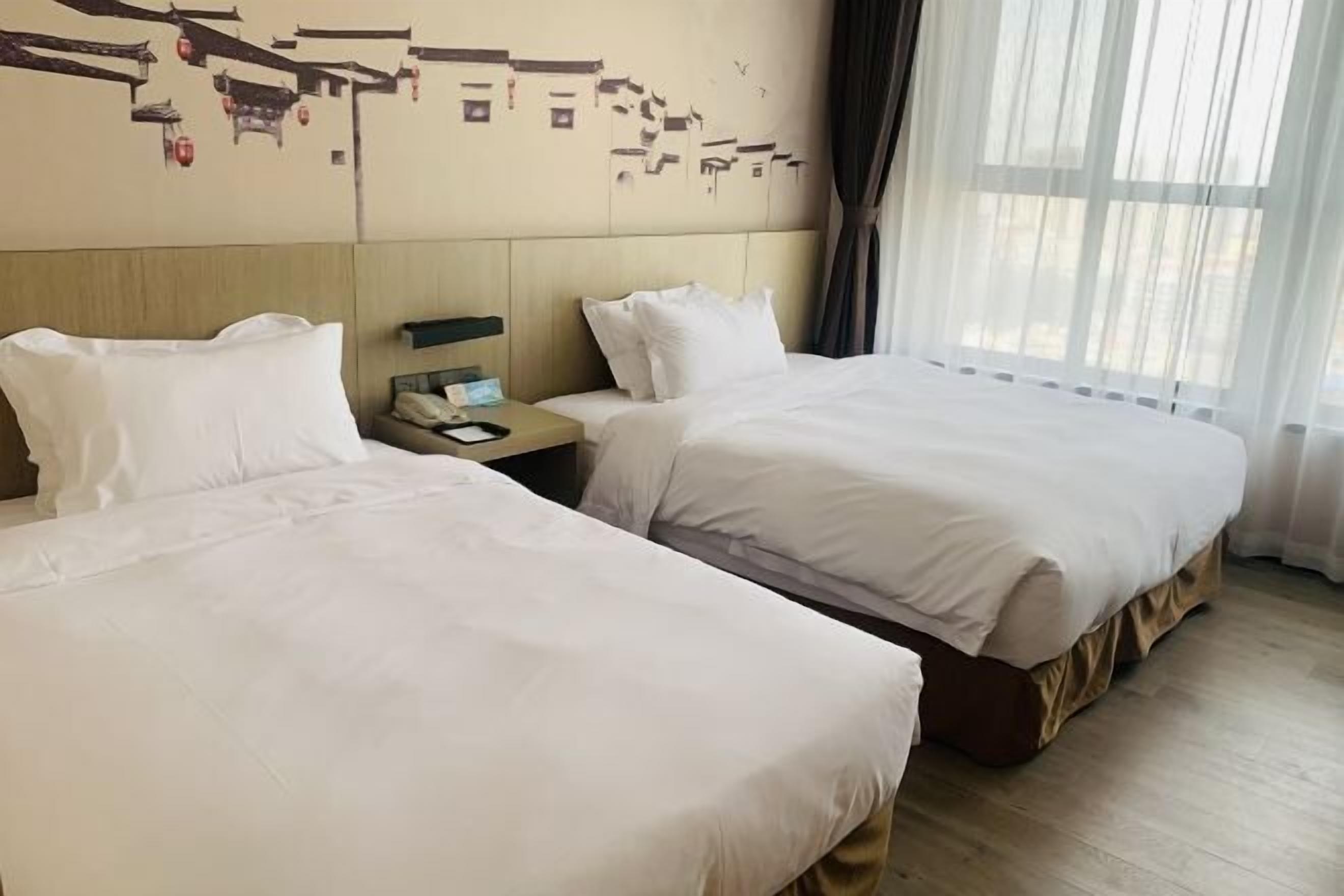 A hotel room with two single beds, a wooden headboard, a nightstand with a phone, and a window with sheer curtains.