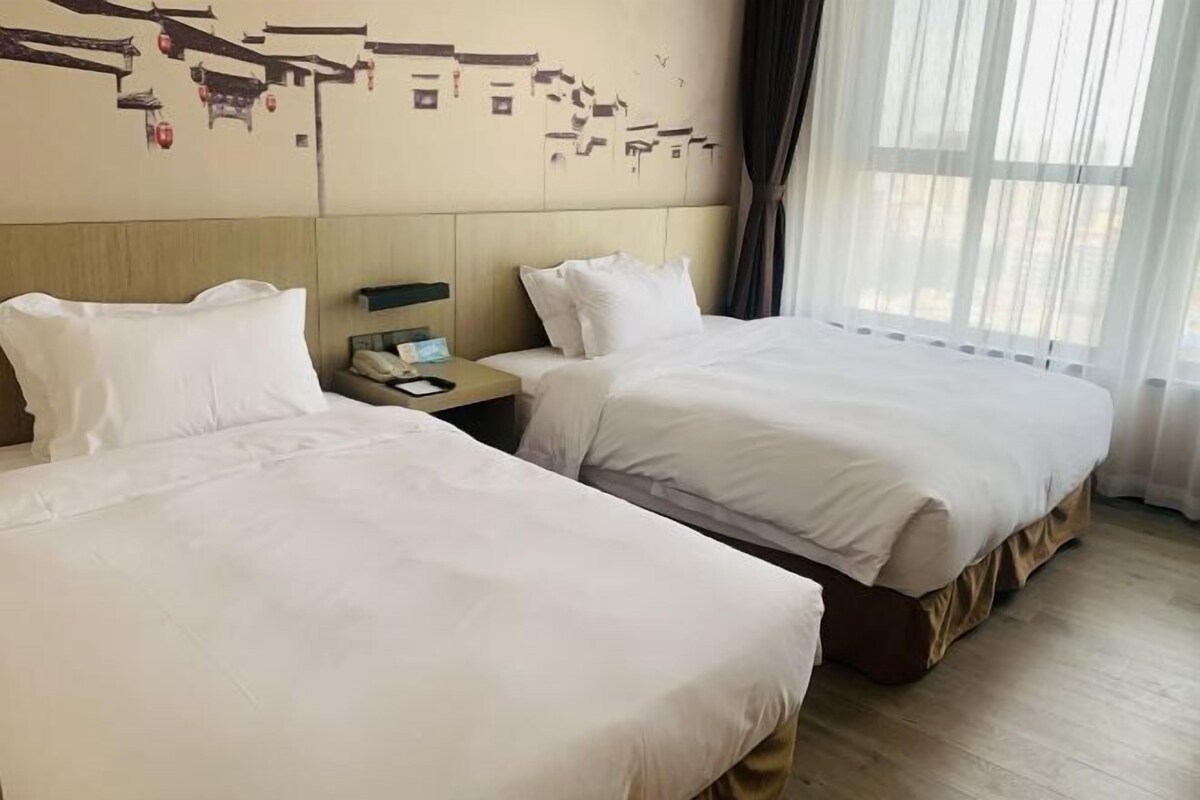 A hotel room with two single beds, a wooden headboard, a nightstand with a phone, and a window with sheer curtains.