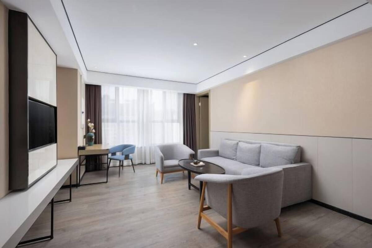 A modern hotel room with a sofa, a dining table with chairs, a flat-screen TV, and a large window with curtains.