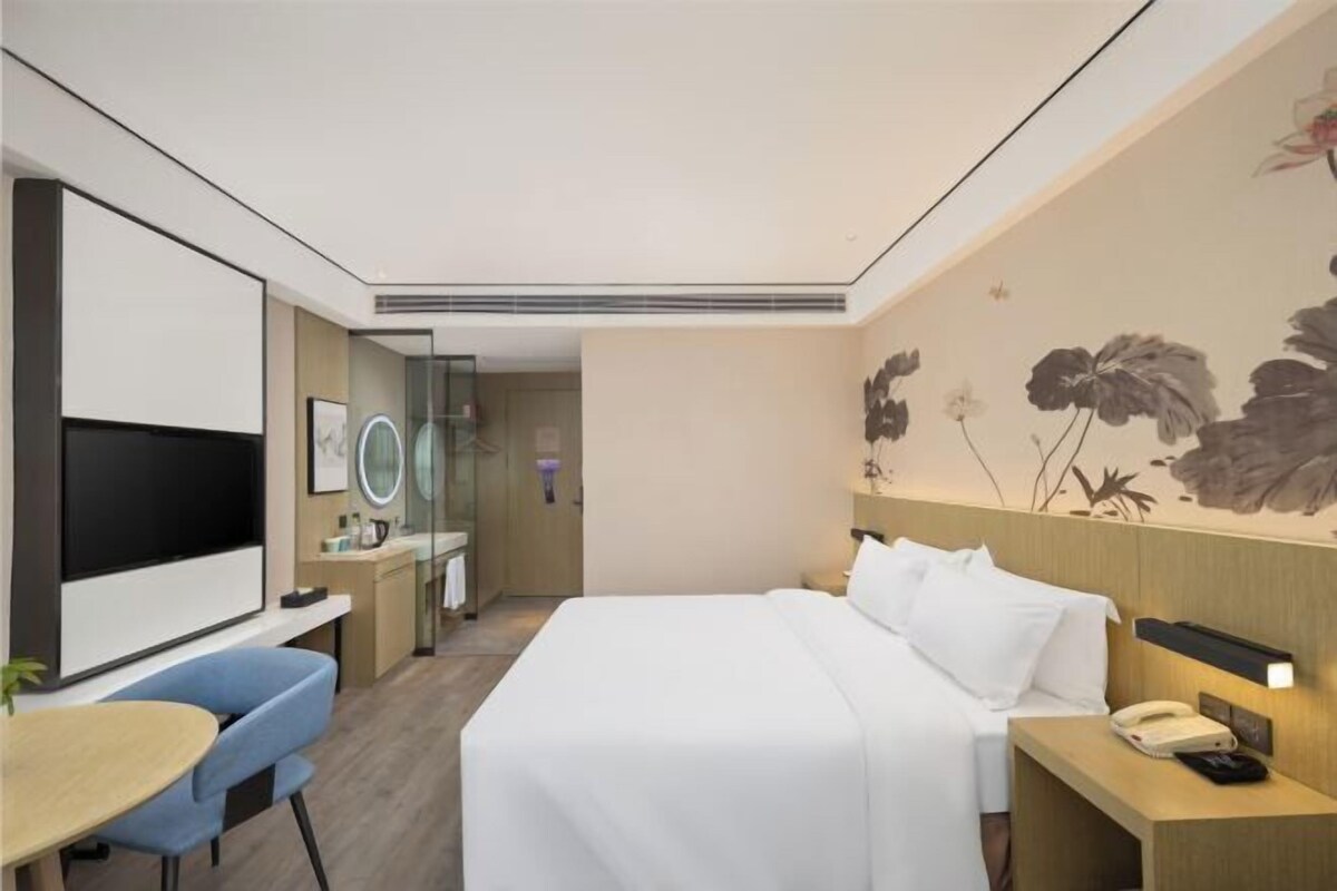 A modern hotel room with a large bed, a desk with a chair, a TV, and a bathroom visible through a glass door.