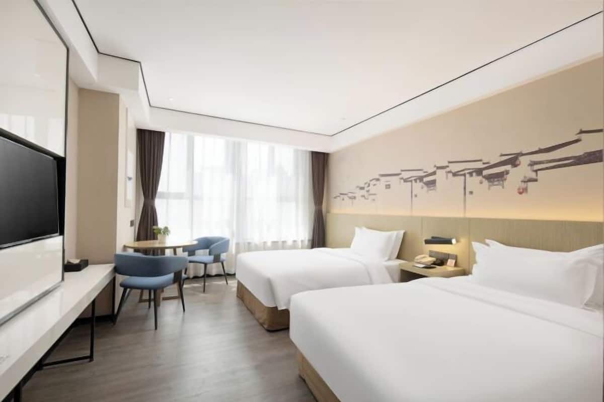 A hotel room with two beds, a TV, a desk, a chair, and a window with curtains.