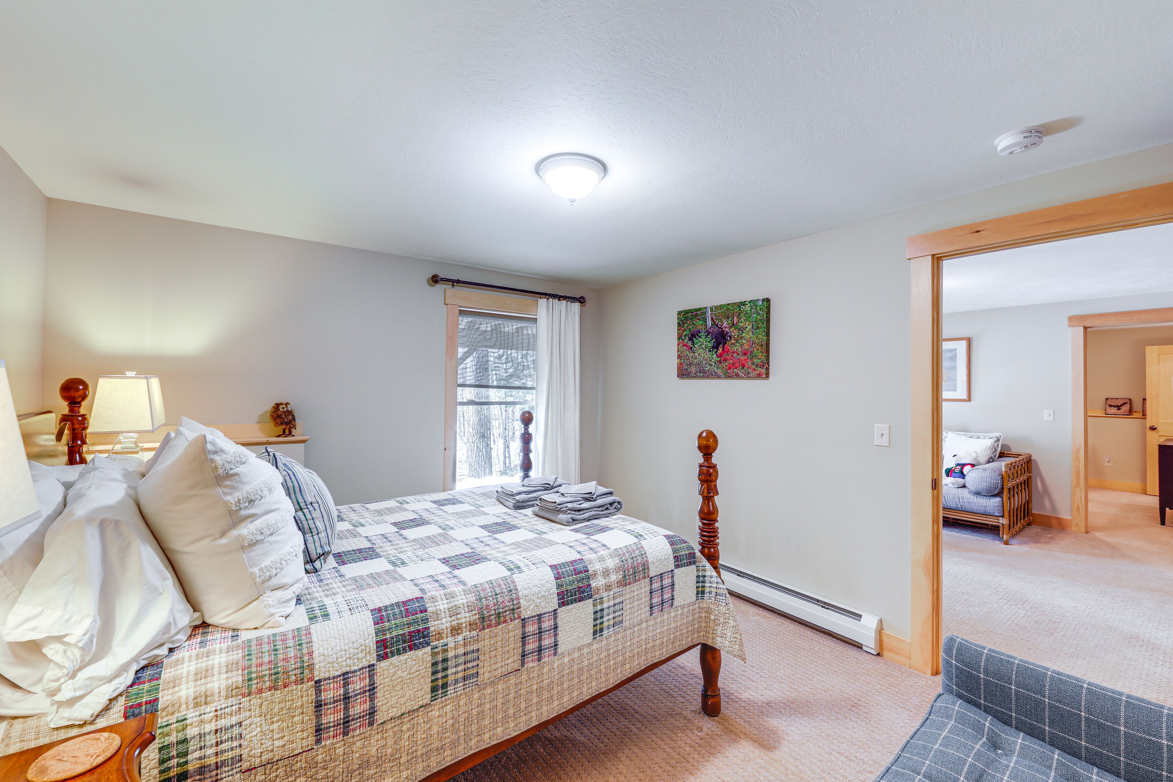 Un dormitorio con cama, mesita de noche, ventana con cortinas, un cuadro en la pared y una puerta que da a otra habitación.