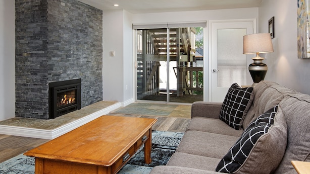 Ein Wohnzimmer mit Kamin aus Stein, einem Holztisch, einem grauen Sofa mit gemusterten Kissen und einer Glasschiebetür zu einem Außenbereich.