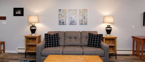 Ein Wohnzimmer mit einem grauen Sofa, zwei Beistelltischen mit Lampen, einem Holztisch als Couchtisch und einem gemusterten Teppich.