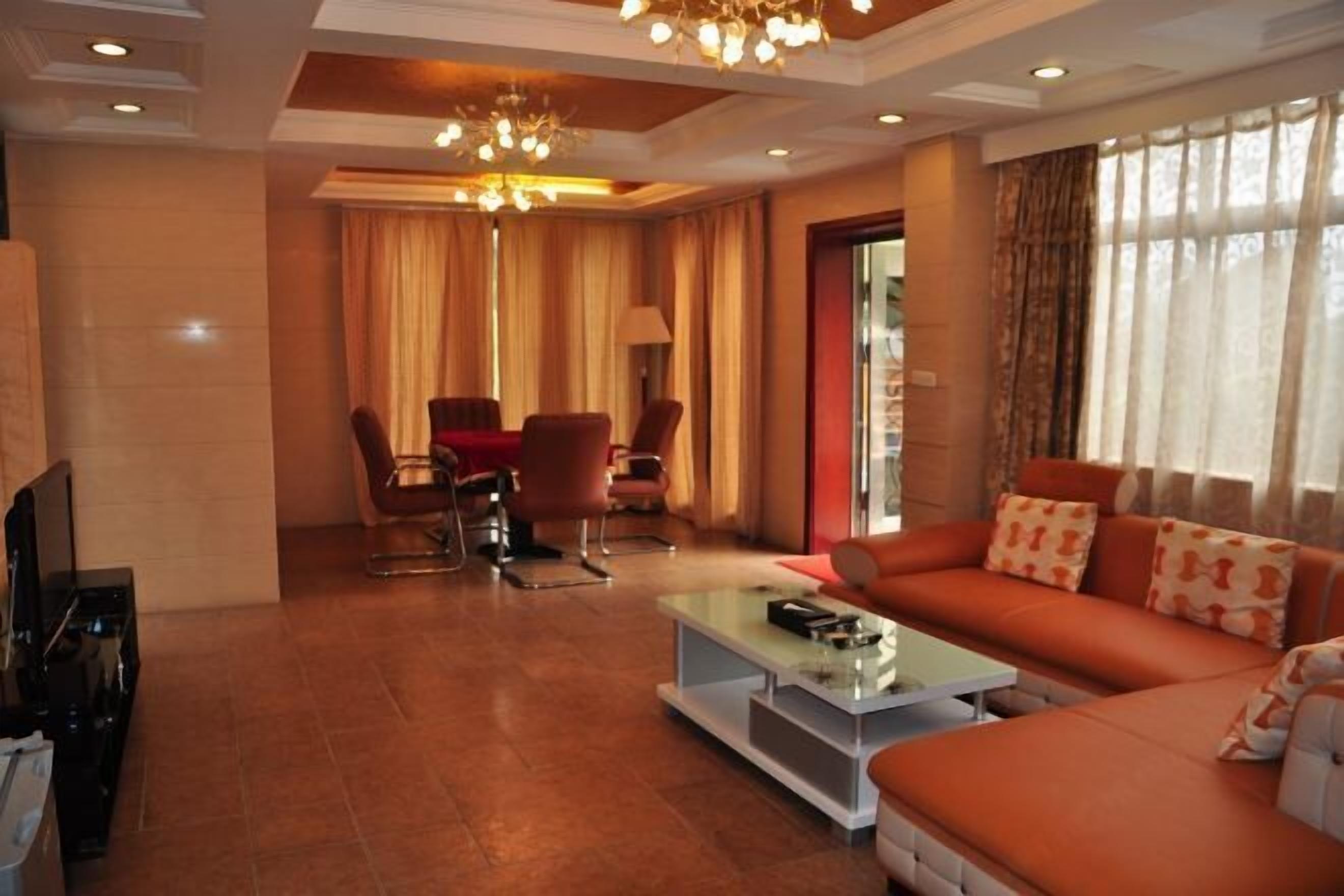 A modern living room with a sofa, coffee table, and chairs. There is a TV on the left and a window with curtains on the right.