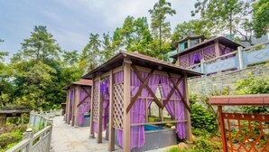 Ein Pfad führt zu einer Reihe aus Holz bestehender Gebäude mit violetten Vorhängen, umgeben von üppigem Grün und Bäumen.