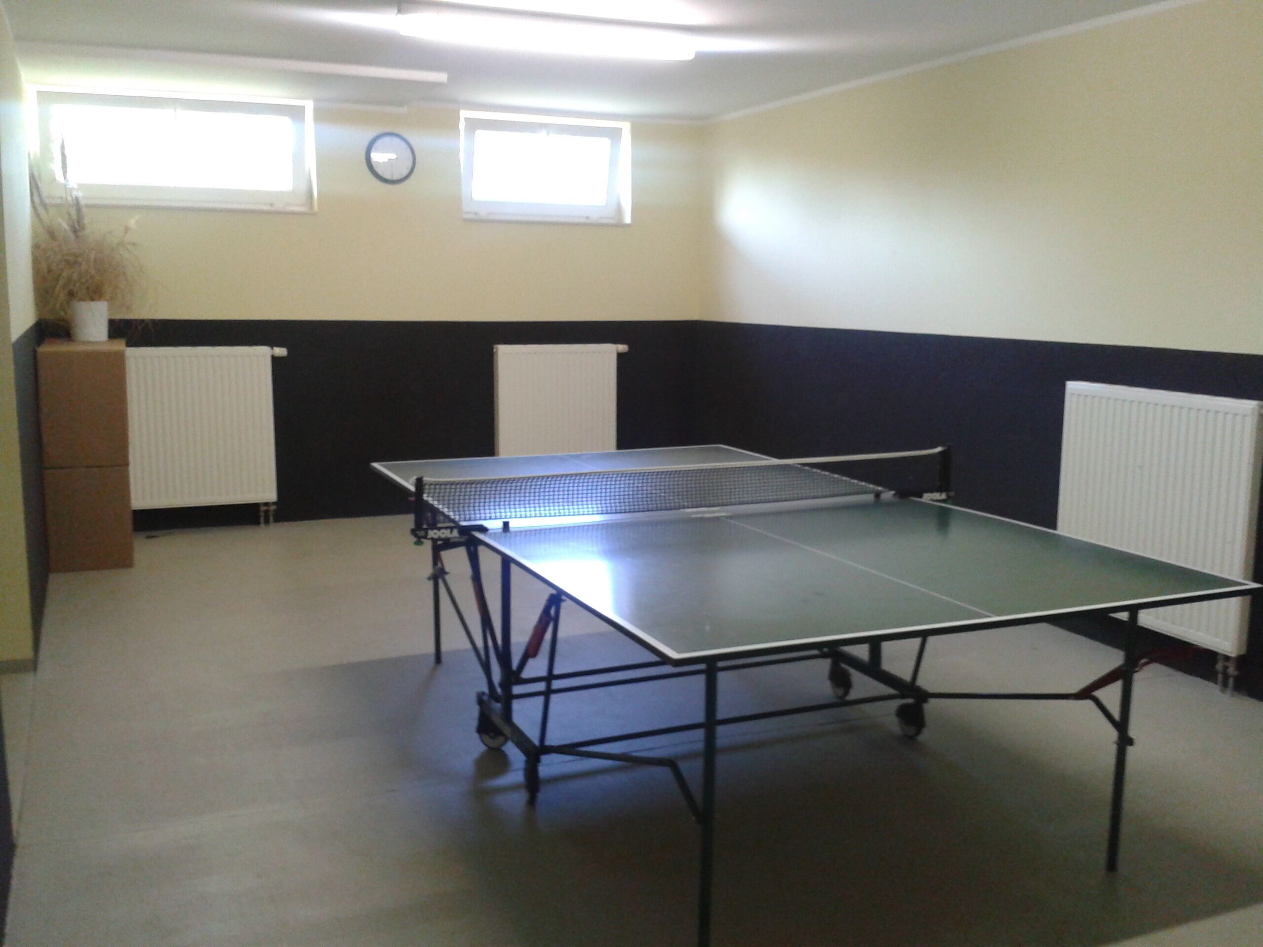 Una habitación con mesa de ping pong, dos radiadores, un reloj y una ventana.