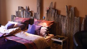 Un dormitorio con cabecera de madera, cama con muchos almohadones, mesita de noche con lámpara y una silla.