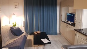 Et lille, godt oplyst værelse med seng, skrivebord, stol, fjernsyn og et tekøkken.